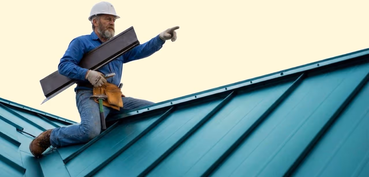 Construction worker in a white hard hat kneeling on a blue metal roof holding a metal sheet and pointing forward.