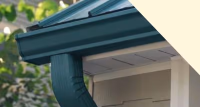 Close-up of a dark green rain gutter and downspout attached to a beige house exterior with blurred trees in the background.