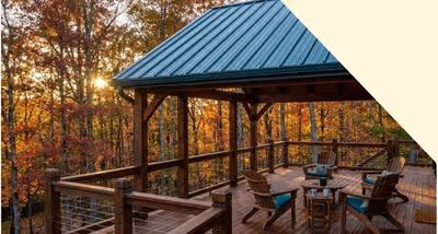 Wooden deck with a metal roof, outdoor furniture, and autumn trees in the background at sunset.