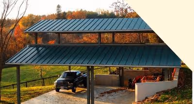 Black pickup truck parked under a green metal roof carport next to some orange construction equipment with autumn trees in the background.