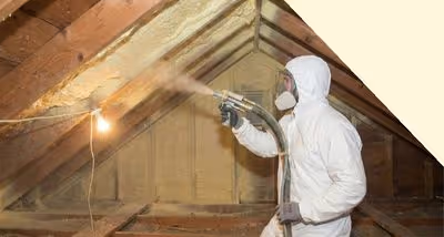 Person in protective suit and mask spraying insulation foam in an attic.