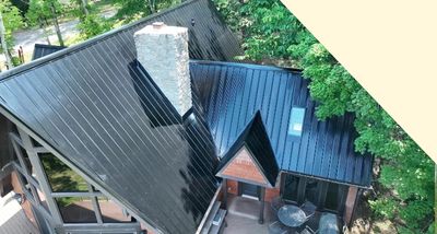 Aerial view of a modern house with a steep black metal roof, stone chimney, and surrounding trees.