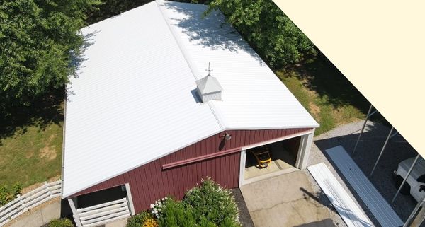 Aerial view of a red barn with a white metal roof, surrounded by trees and greenery.