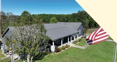 Large single-story gray building with a covered porch, surrounded by green grass and trees, under a clear blue sky.