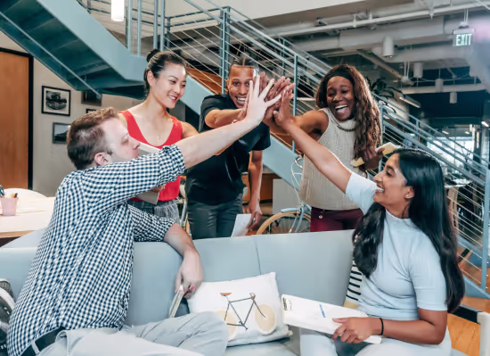 Diverse group of five people sitting and standing in a modern office, smiling and giving a group high-five.