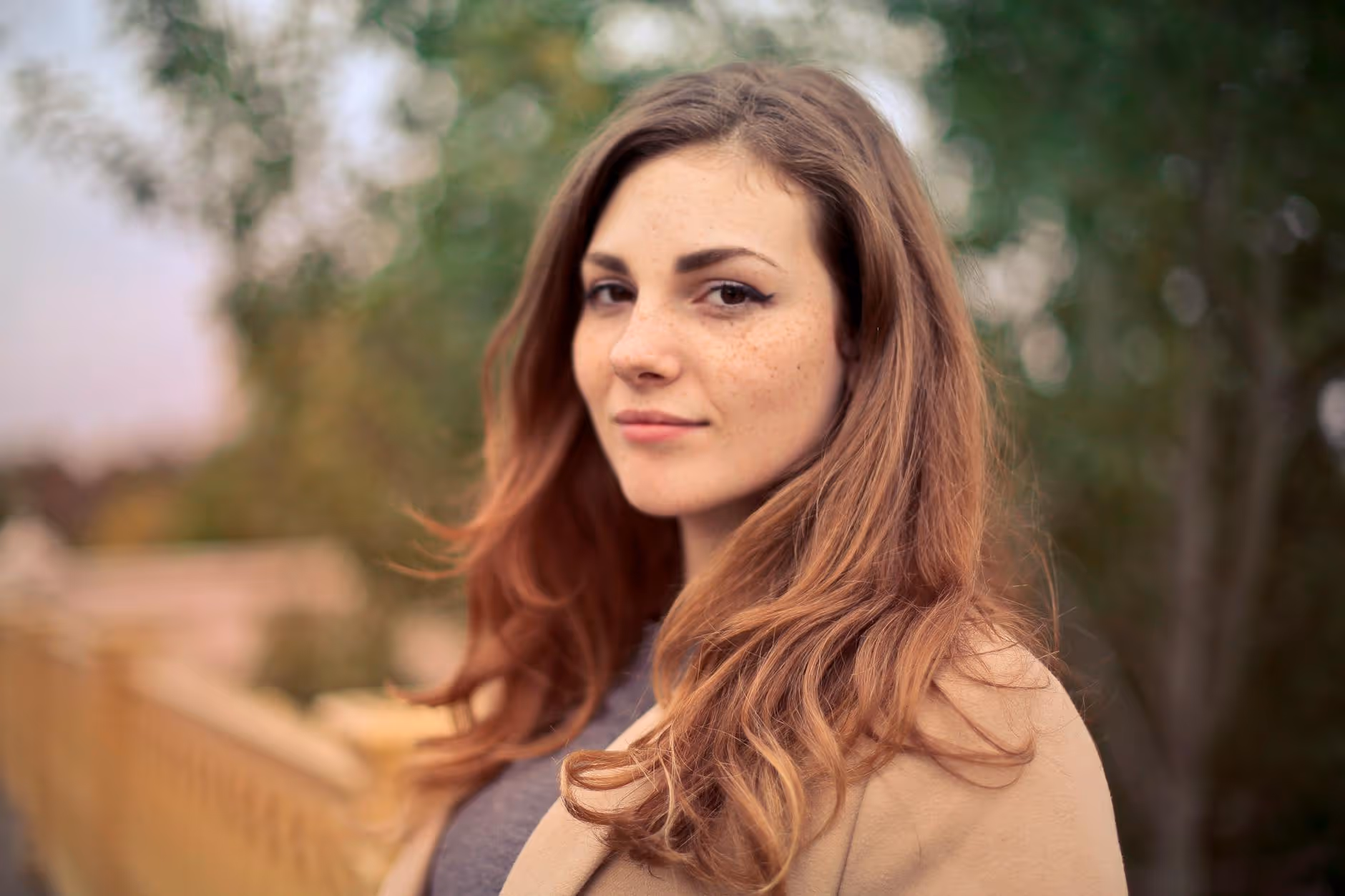 Young woman with long light brown hair and freckles looking at the camera outdoors with blurred trees and railing in the background.