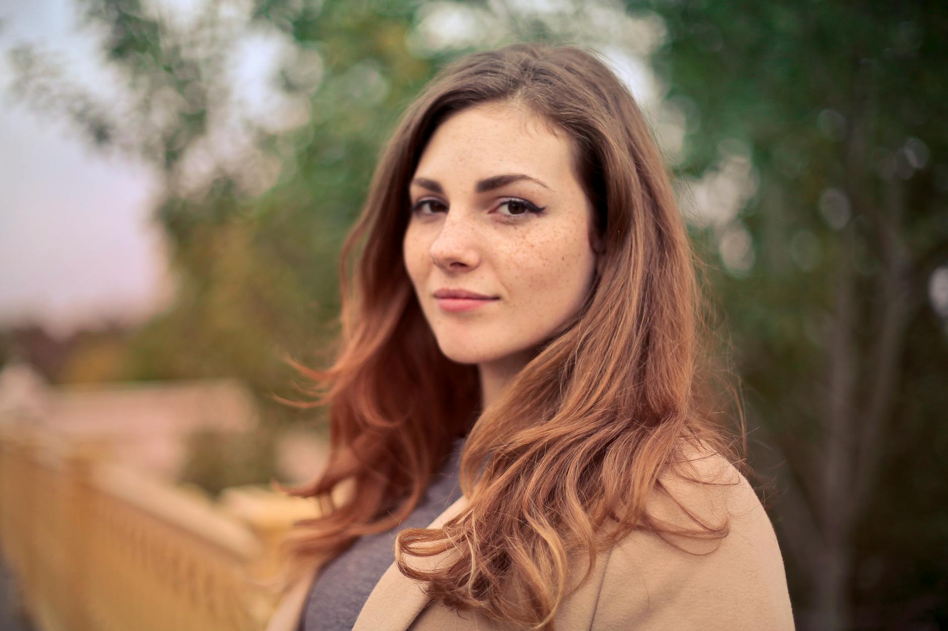 Young woman with long light brown hair and freckles looking at the camera outdoors with blurred trees and railing in the background.
