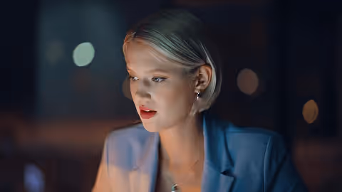 Woman with short blonde hair wearing a blue blazer looking at a lit screen in a dark room.