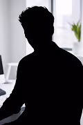 Silhouette of a man sitting indoors by a window with a blurred plant in the background.