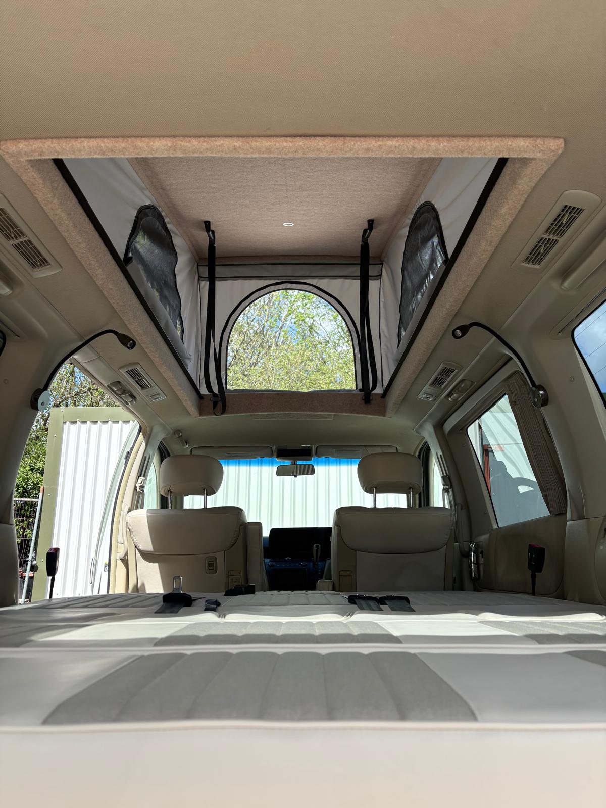 Interior of a camper van showing rear seats, extended bed, and a pop-up tent roof with a window.