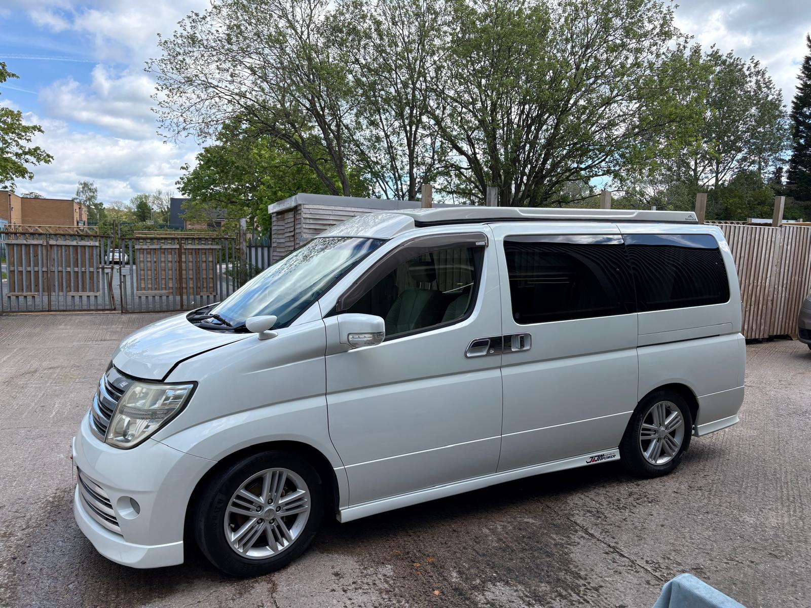 White passenger van parked on concrete with trees and wooden fence in the background.