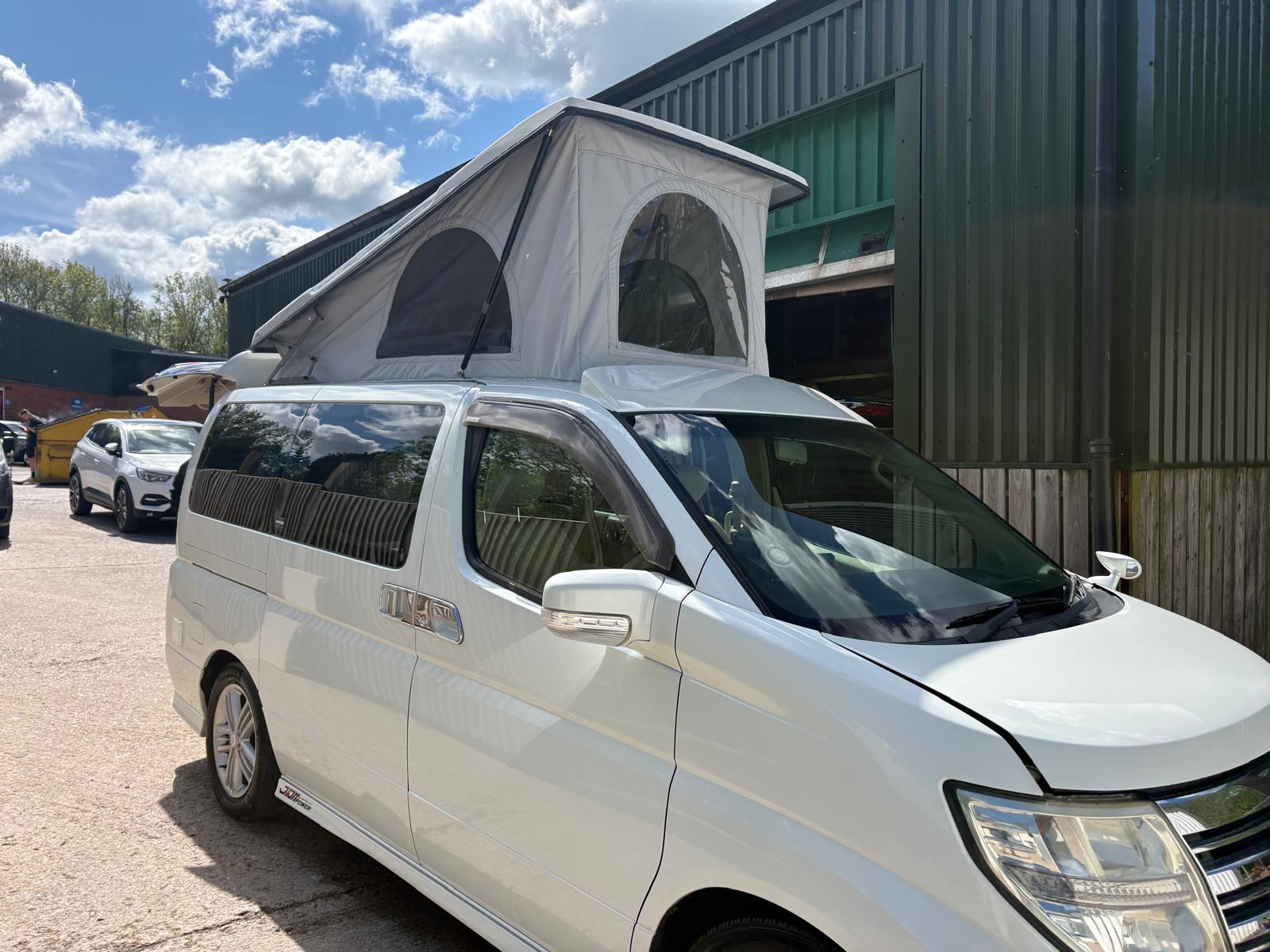 White camper van with roof tent parked beside a green industrial building under a partly cloudy sky.