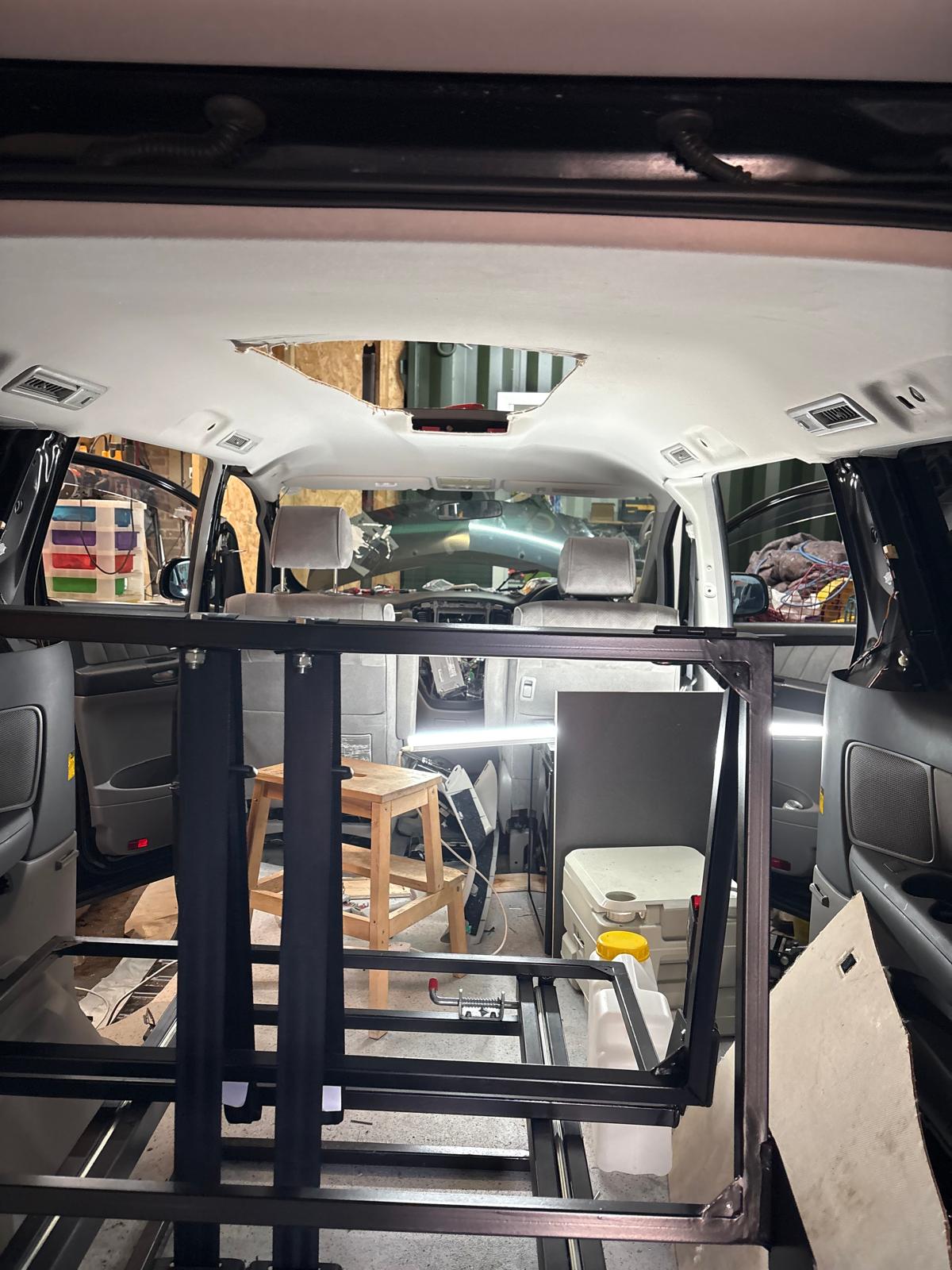 Interior of a vehicle with seats removed, showing metal frame, wooden stool, cooler, and tools inside.