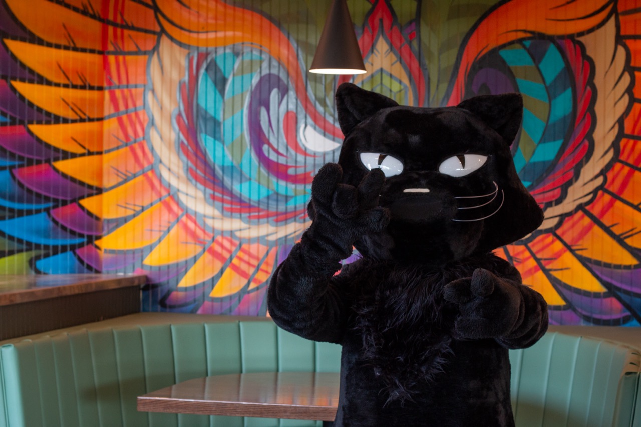 Person in black cat costume posing with one hand making a rock sign and the other pointing forward, standing in front of a colorful mural of wings.