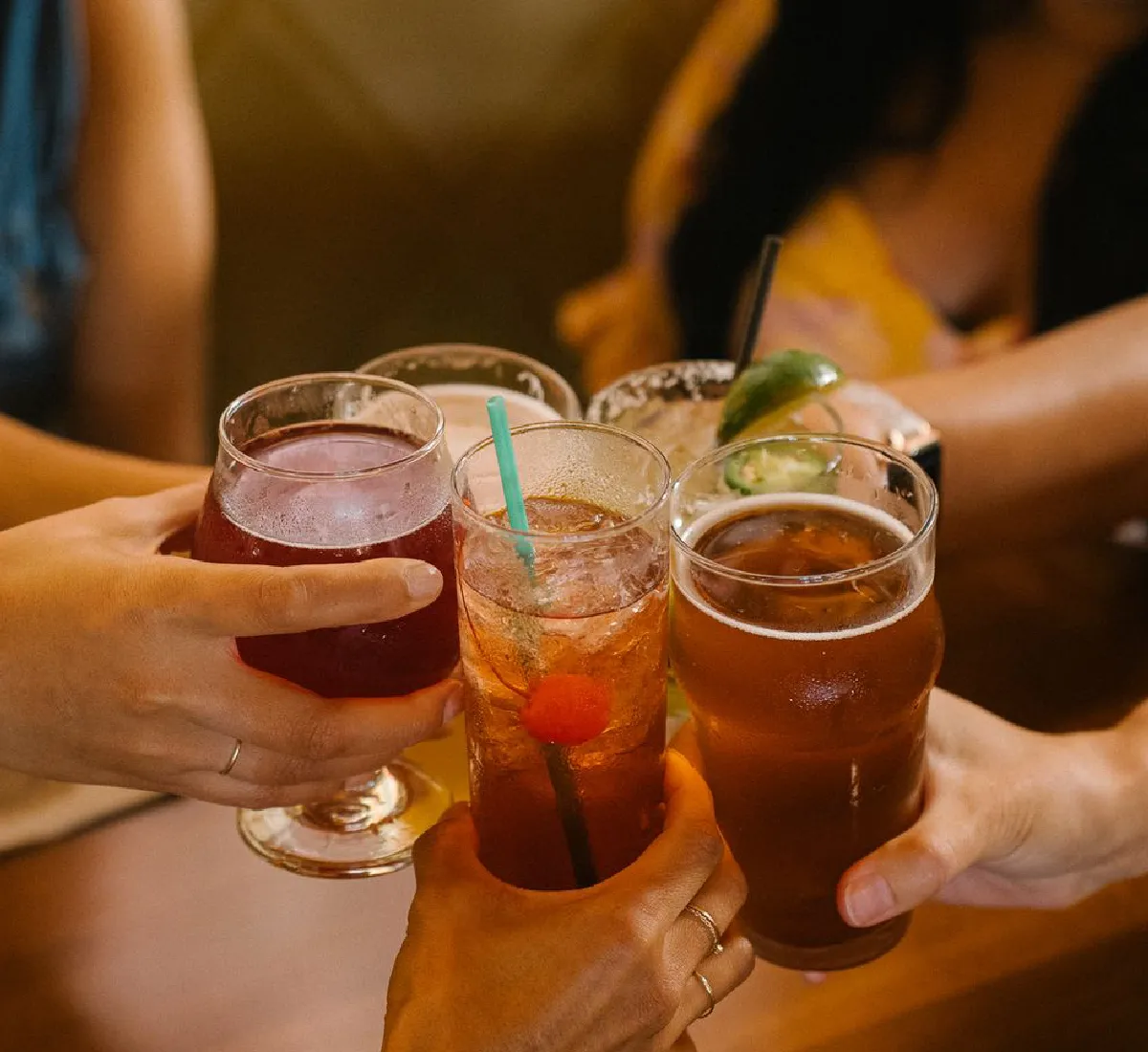 Four hands holding different drinks — a red cocktail, a tall iced drink with a cherry and straw, a pint of beer, and a margarita with a lime wedge — as they toast together.