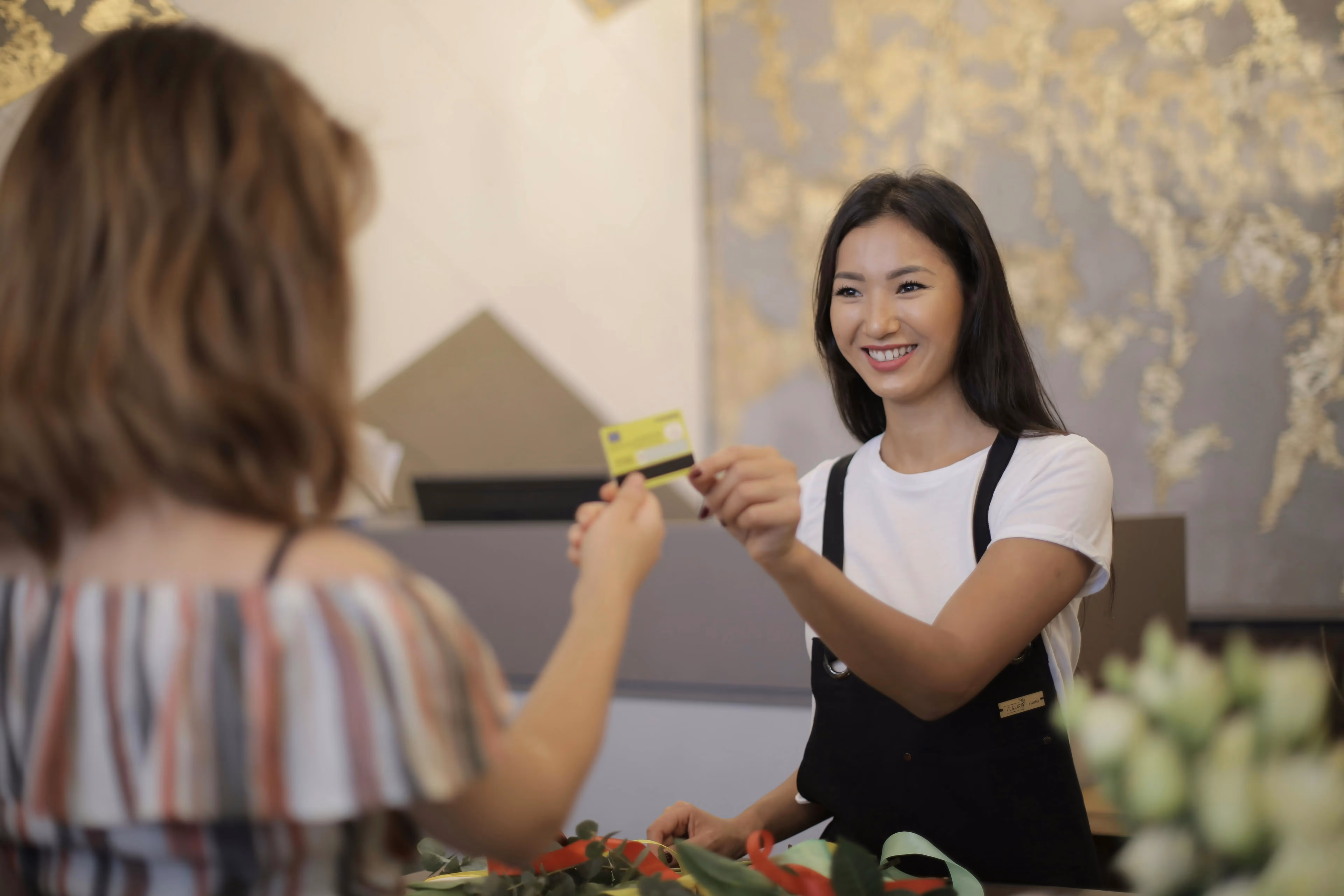 Woman passing a credit card to another woman, both smiling, in a simple setting.