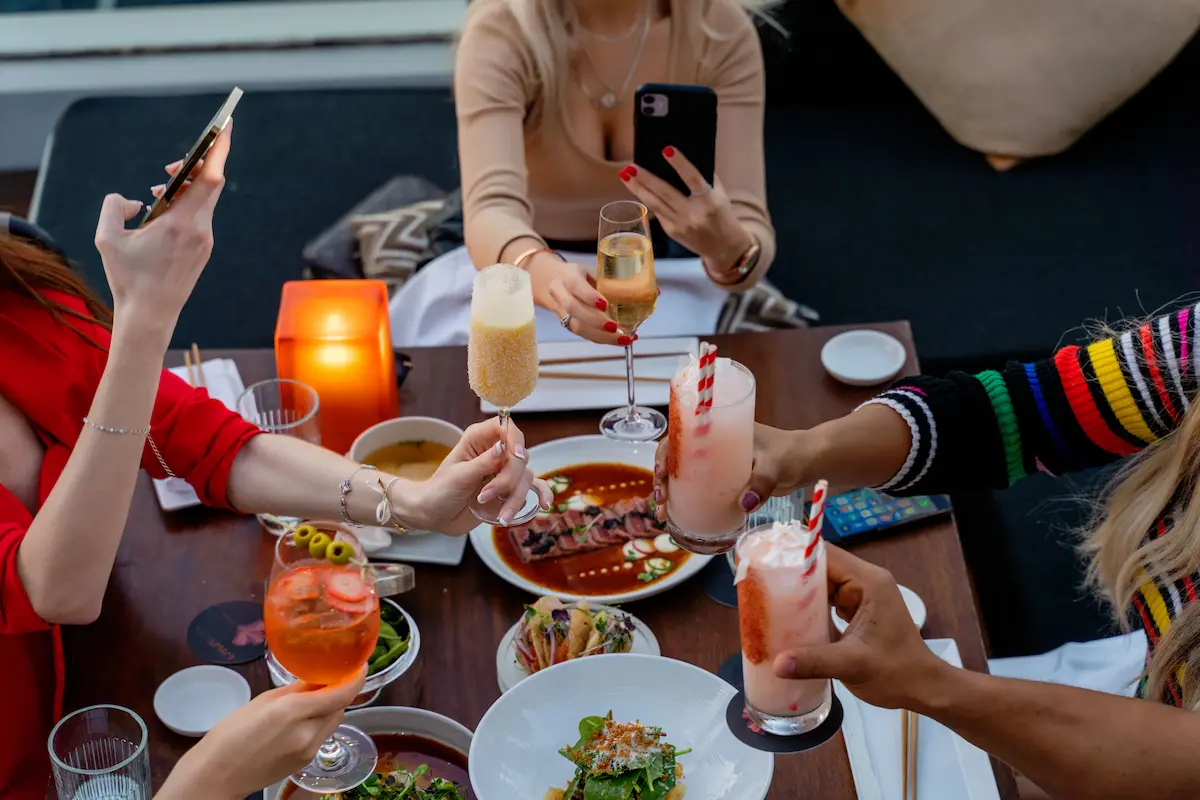 Group of friends clinking drinks at a restaurant table with food, representing dining out with Amex Resy credit card benefits
