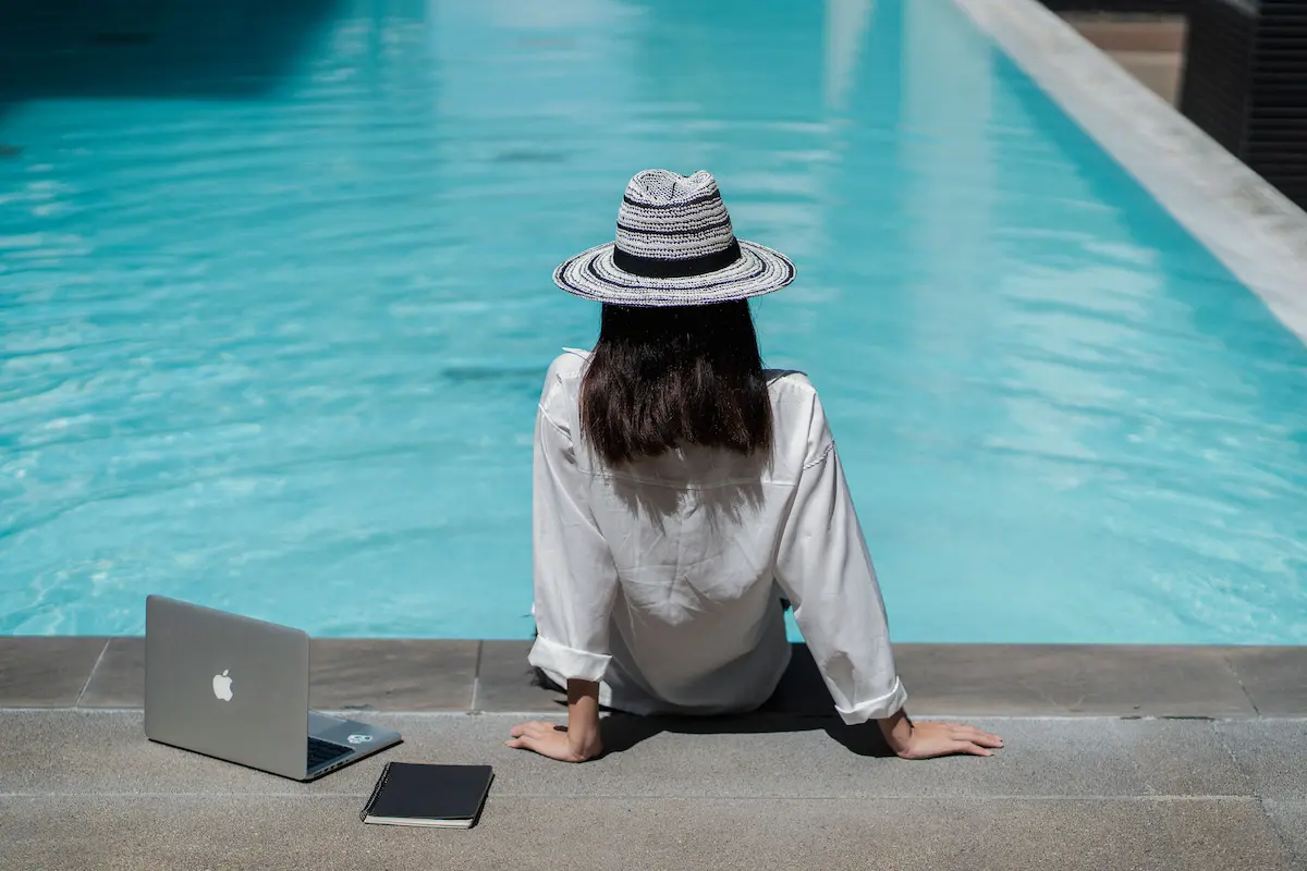 Woman in a sun hat sitting poolside with a laptop and notebook beside her at a resort