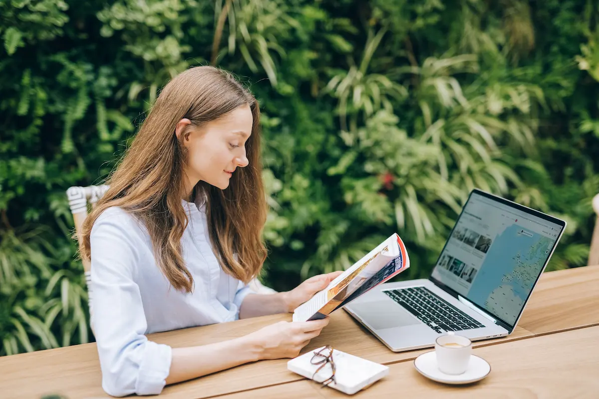 Woman reading a travel magazine at an outdoor table with a laptop showing a map and travel booking site open beside her