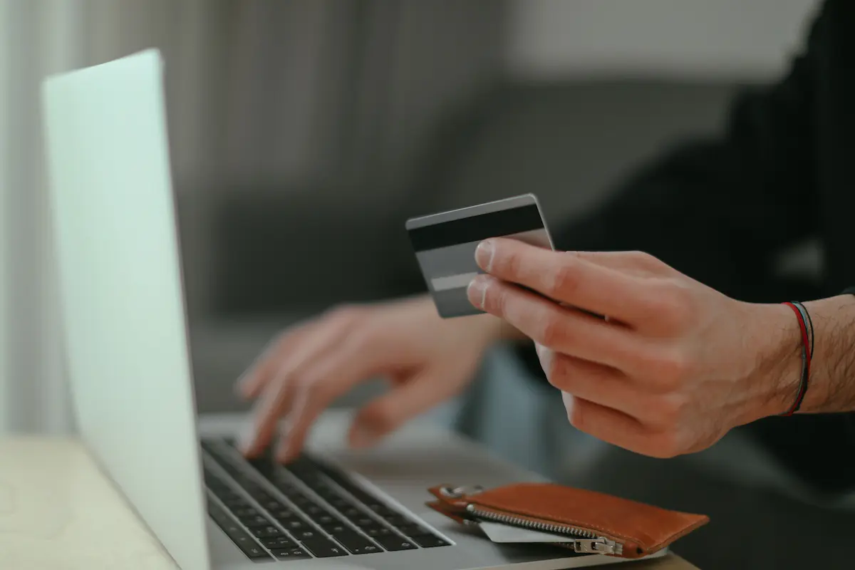 Close-up of a hand holding a credit card while typing on a laptop keyboard, with a leather wallet resting nearby