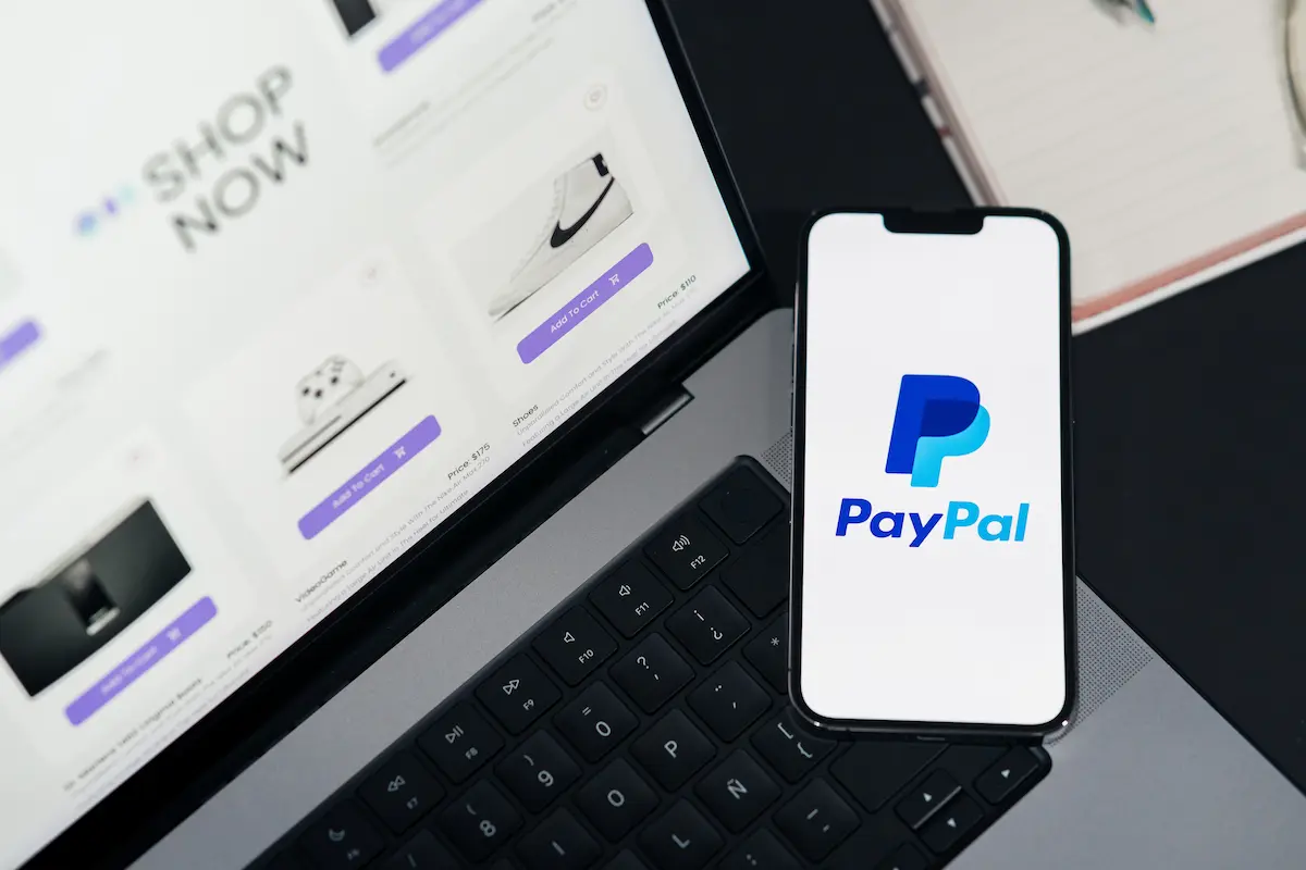 Smartphone displaying the PayPal logo next to a laptop showing an online shopping cart page