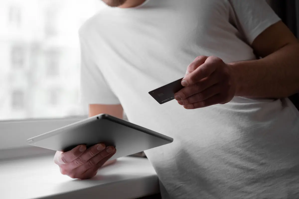 Man in a white t-shirt holding a dark credit card in one hand and a tablet in the other, preparing to enter payment information