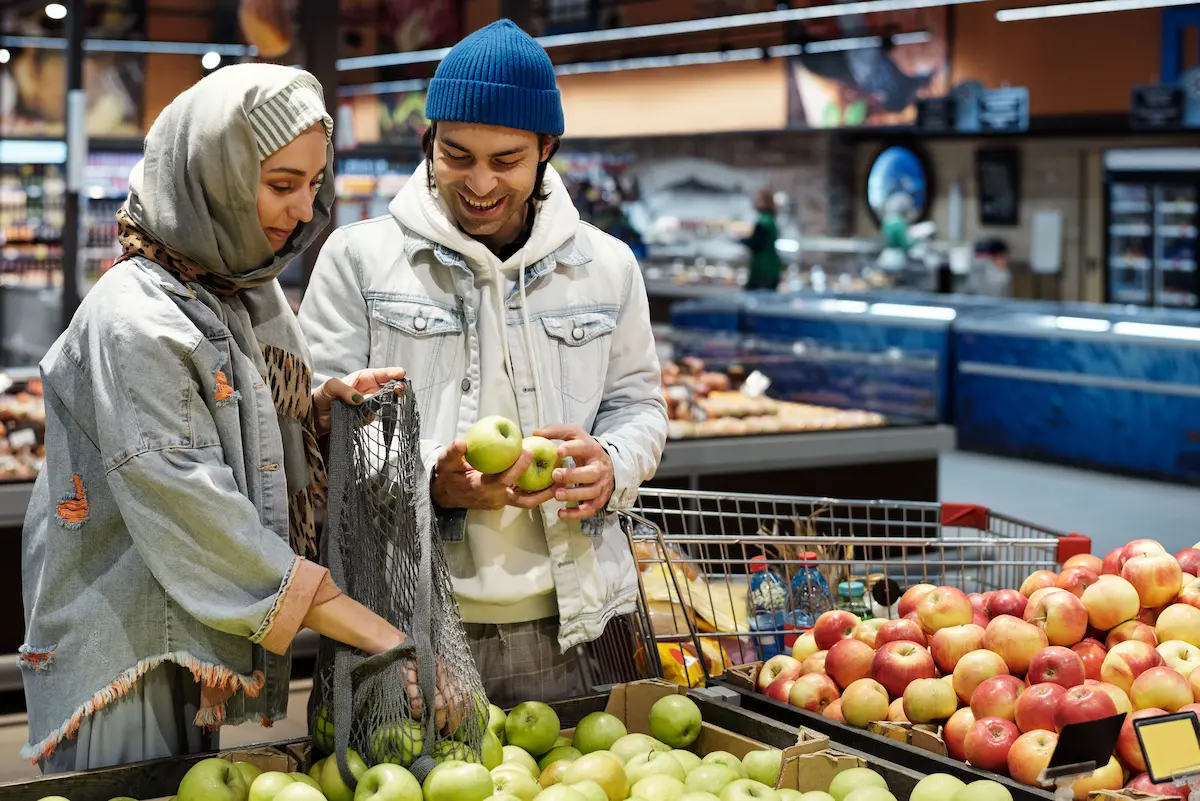 Two shoppers picking out apples in the grocery store produce section, representing everyday grocery spending covered by cash back credit cards.
