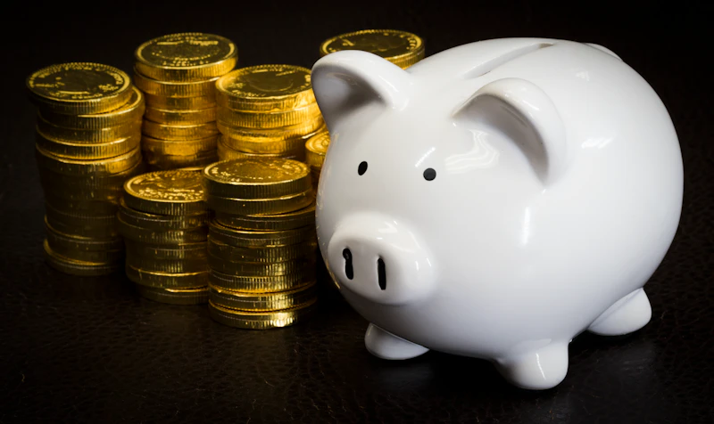 A white ceramic piggy bank beside stacks of gold coins on a dark surface