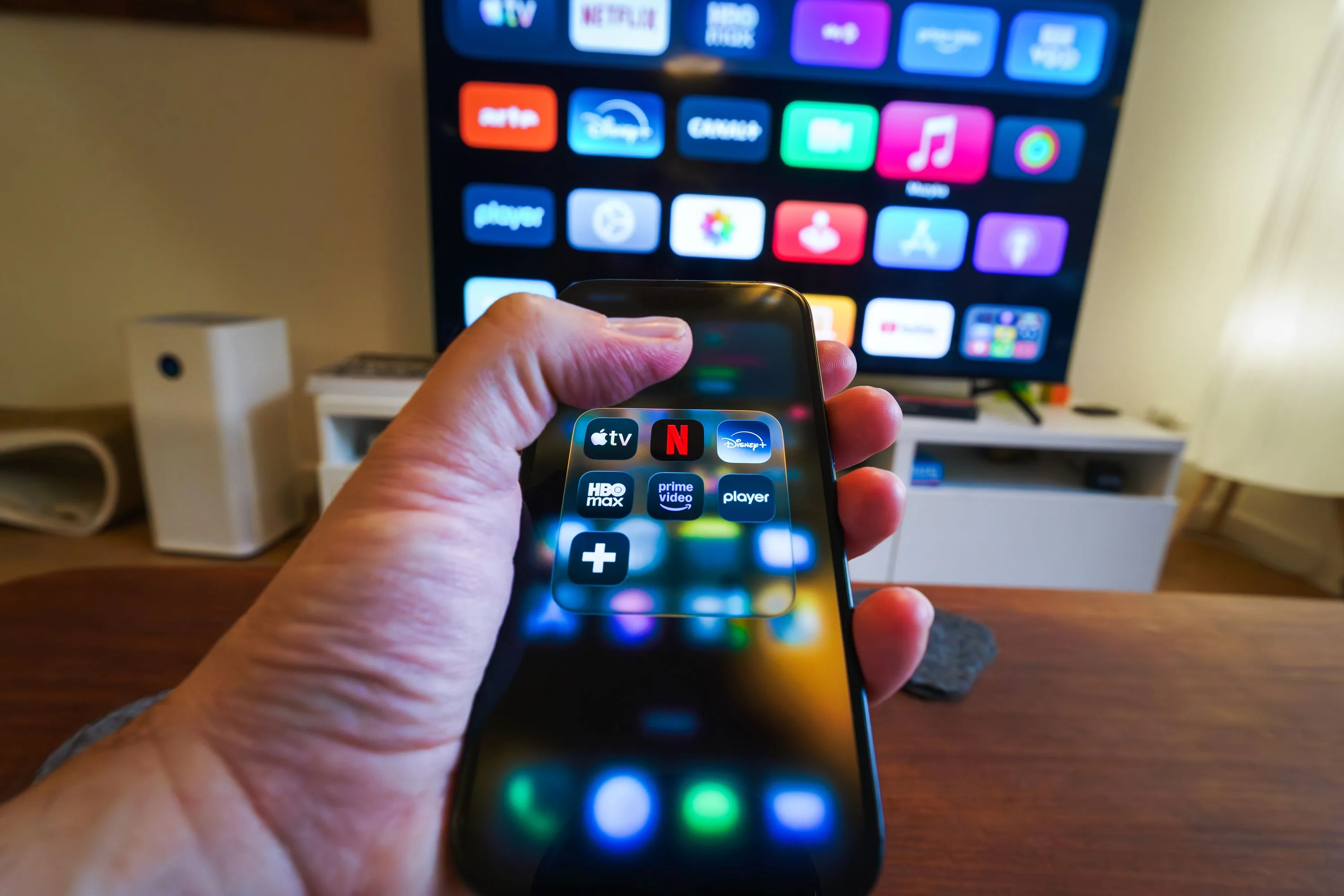 Hand holding a phone with a streaming app switcher showing Apple TV, Netflix, Disney+, HBO Max, and Prime Video in front of a smart TV home screen