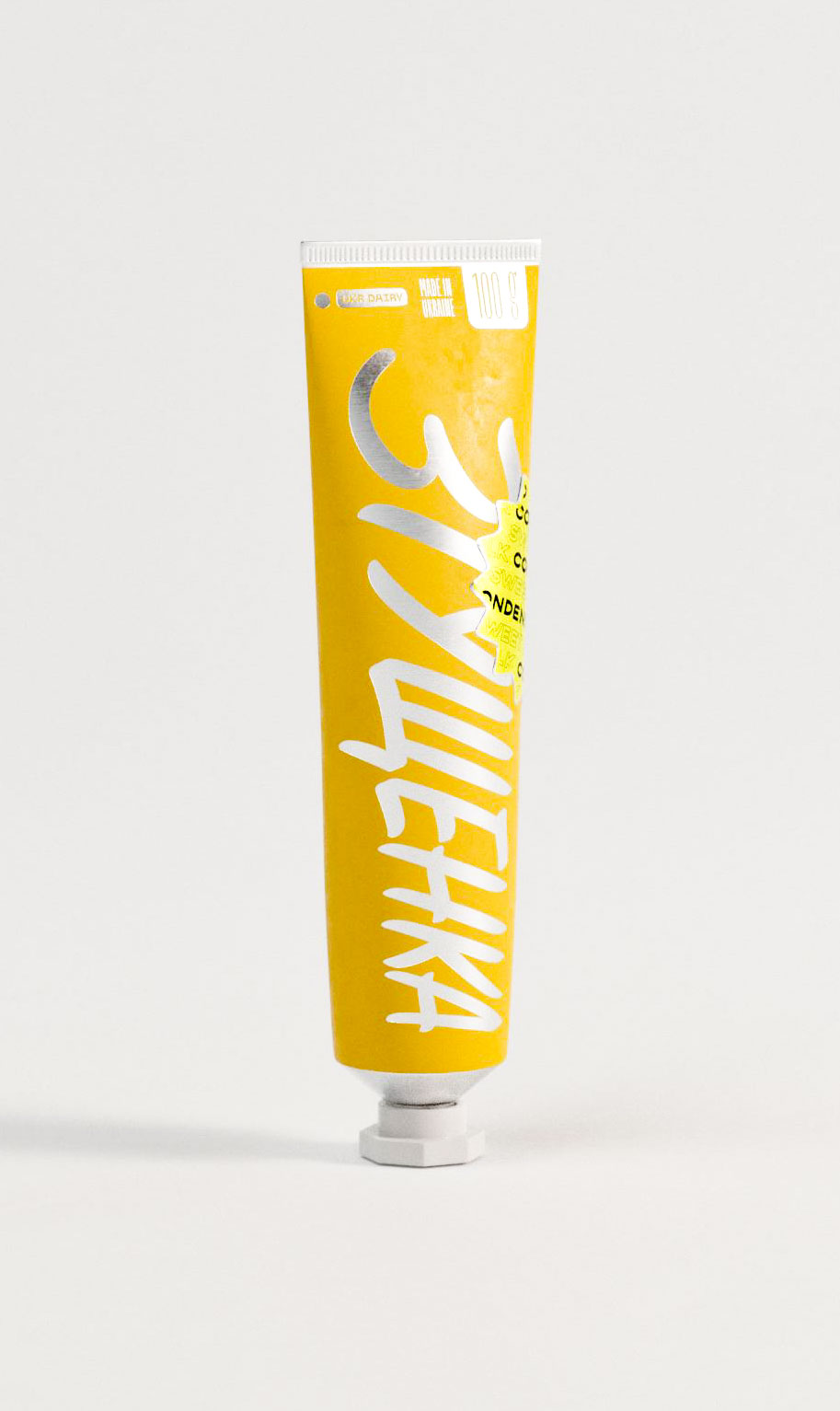 A tube of condensed milk is photographed upright against a clean, white background. The packaging is designed in a vivid yellow hue, featuring a large handwritten-style wordmark in white Cyrillic lettering stretching vertically across the body. A small silver cap grounds the base. Minimal product information sits at the top, including weight, origin ("Made in Ukraine"), and a neon starburst badge with layered “CONDENSED” text. The graphic design approach uses expressive typography, contrast, and nontraditional layout, demonstrating how a packaging design agency reimagines food staples with cultural energy and visual punch.