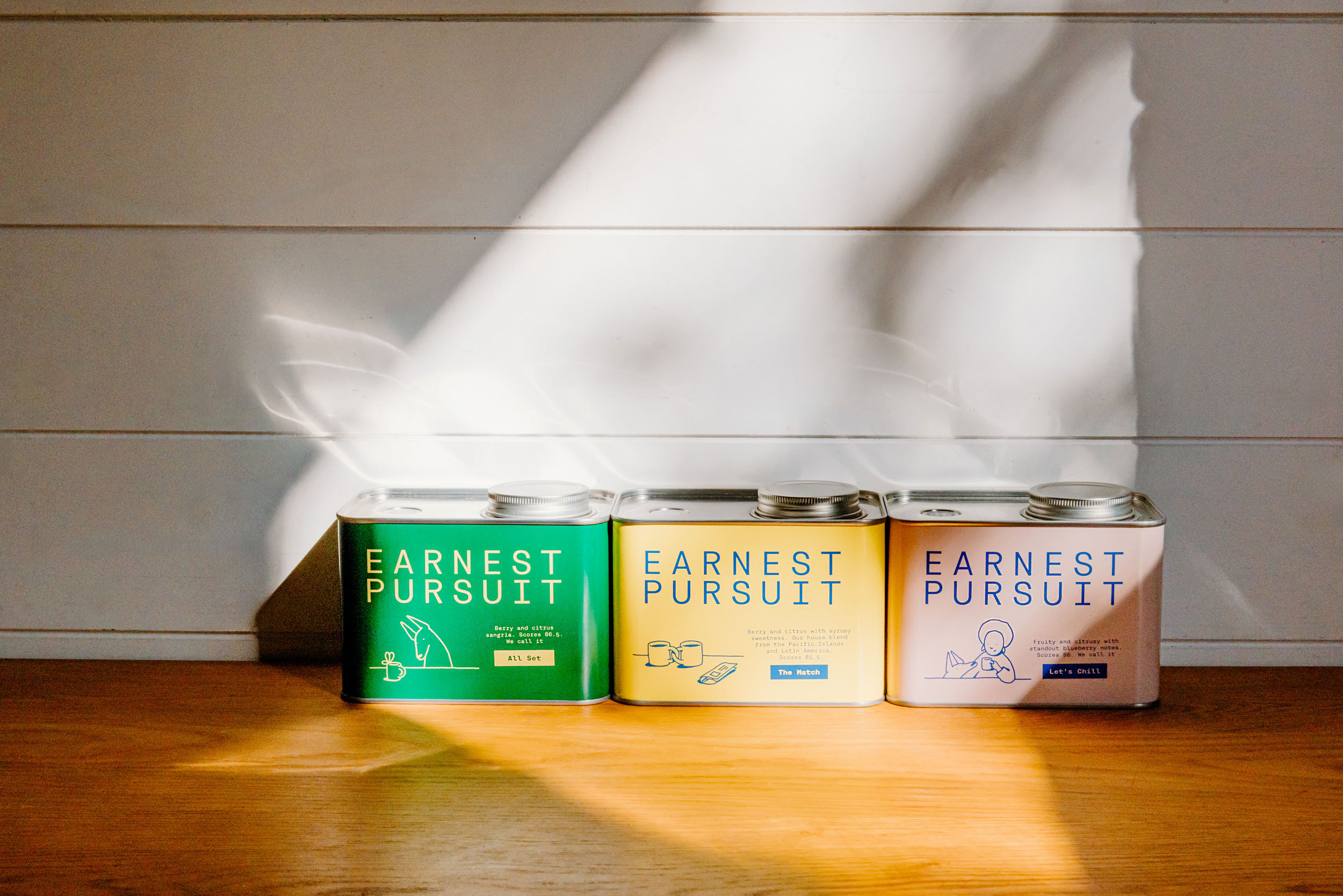 Adeline Artistry photographs three Earnest Pursuit tins in a row on a wood counter against white shiplap, a shaft of sunlight angling across the boards. Green "All Set", yellow "The Match" and peach "Let's Chill" each carry the wordmark, a character drawing and a tasting line, the yellow one reading "Berry and citrus with syrupy sweetness. Our house blend from the Pacific Islands and Latin America." Daylight warms the yellow toward cream and the peach toward pink, a reminder that printed color is only as stable as the window. The packaging design for all three is by Art of Packaging.