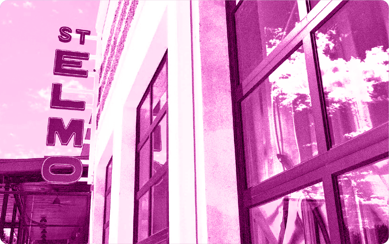 Vertical storefront sign reading 'ST ELMO' mounted on a white building with large glass windows reflecting trees and sky.