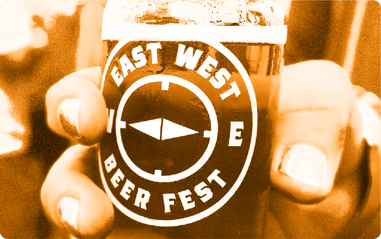 Close-up of a hand holding a beer glass with the logo 'East West Beer Fest' featuring a compass design.