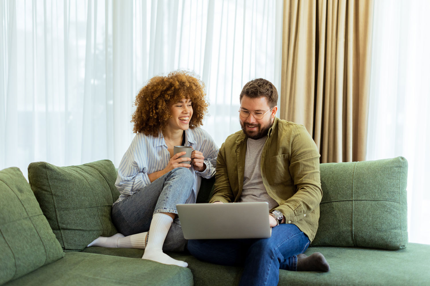 Smiling couple sitting on a green couch, looking at a laptop together, with the woman holding a mug.
