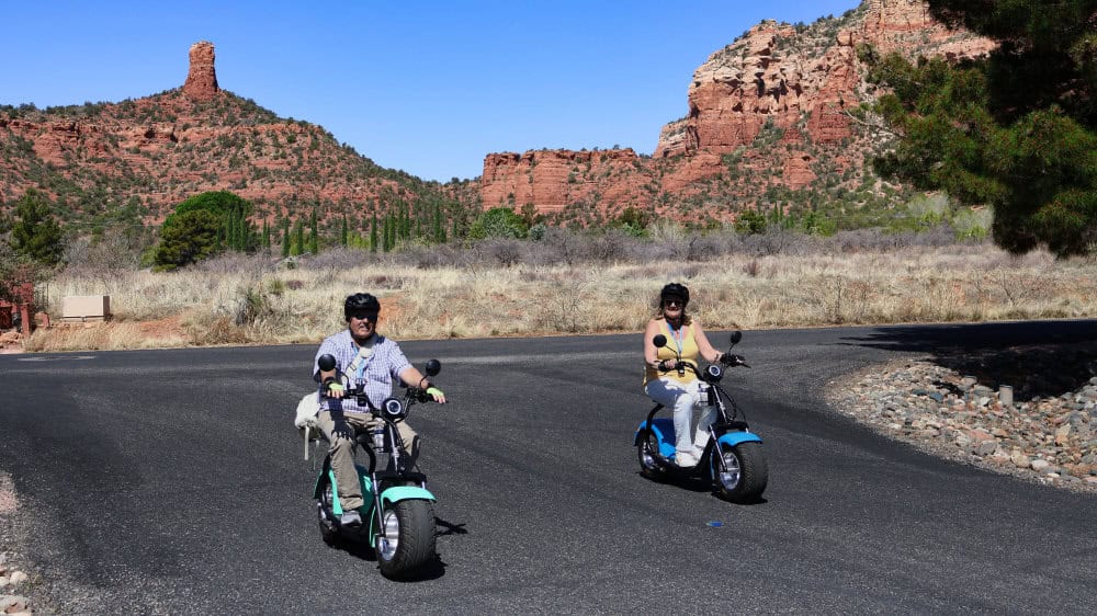 couple on escooters riding on sedon road with red rock mountains behind them