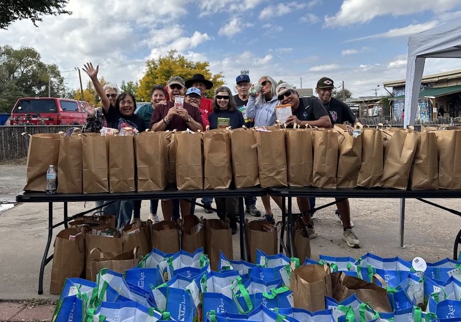 A smiling young girl in a tie-dye tank top carrtying a brown cardboard to-go container with fresh tomatoes and cheese wrapped in plastic stacked on top