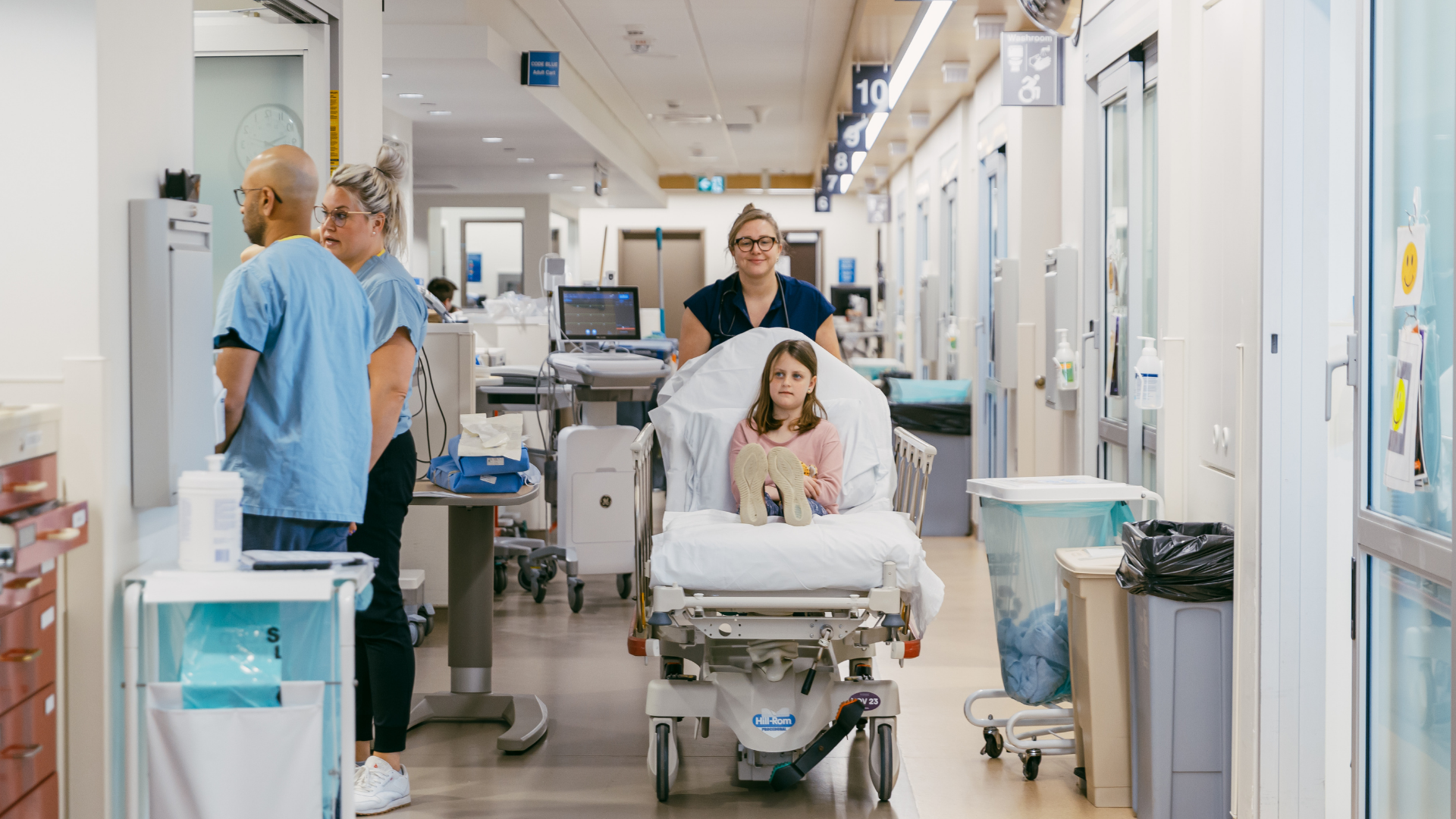 Male doctor using a stethoscope to listen to the chest of a young girl sitting on a hospital bed in a medical examination room.