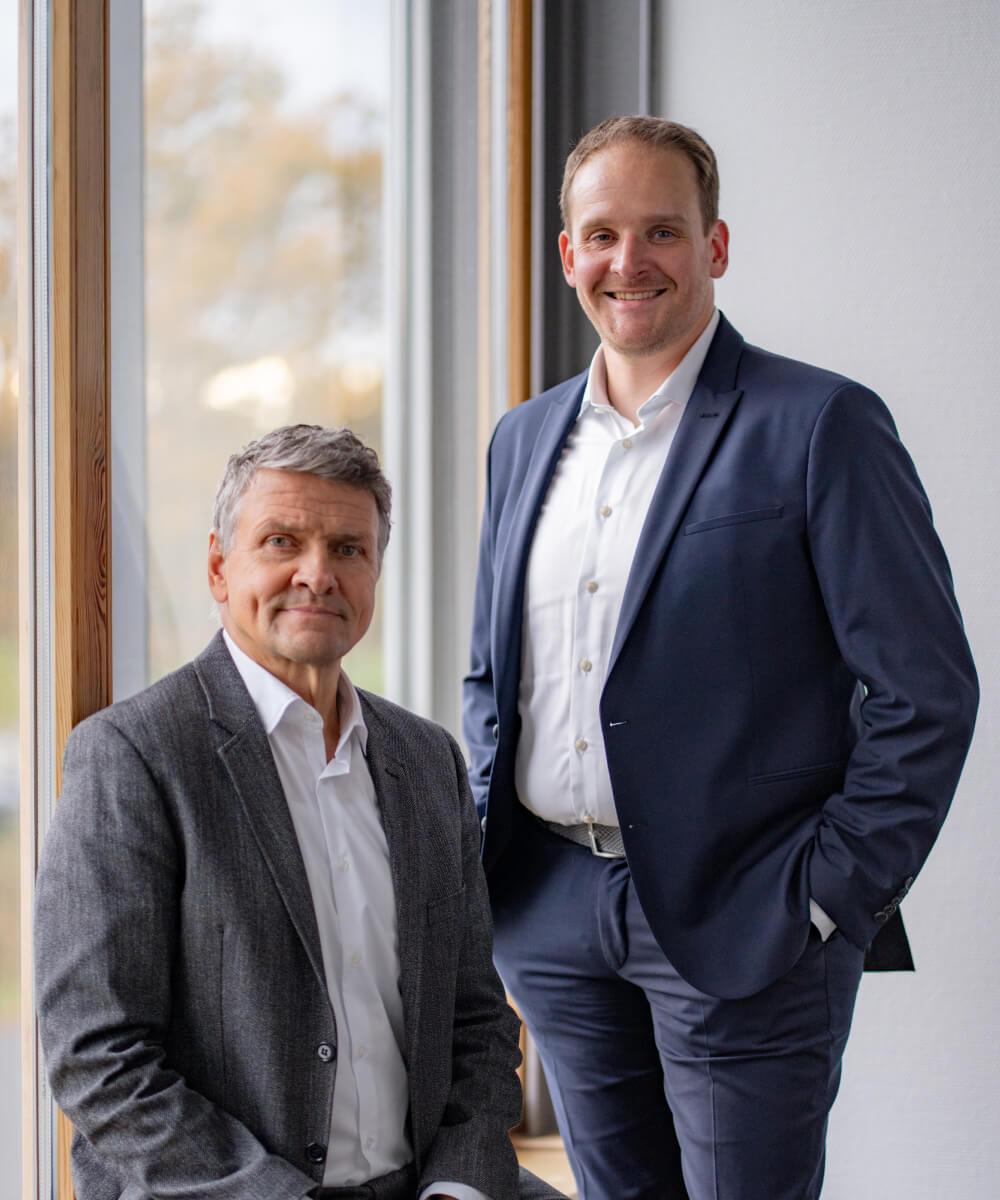 Die beiden rde-Geschäftsführer stehen nebeneinander vor einem Fenster. Links Detlev Falkner mit grauem Sakko, rechts Felix Schürmann mit dunkelblauem Anzug und weißem Hemd.
