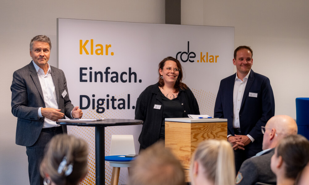 Detlev Falkner, Jasmin Peppel und Felix Schürmann stehen vor dem Publikum der RDE Kundentage. Hinter ihnen steht ein Banner mit der Aufschrift "rde.klar. Klar. Einfach. Digital."