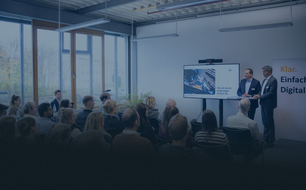 Felix Schürmann und Detlev Falkner stehen vor dem Publikum. Am Bildschirm hinter ihnen stehen die Worte: "Warum rde Ihr langfristiger Partner ist."
