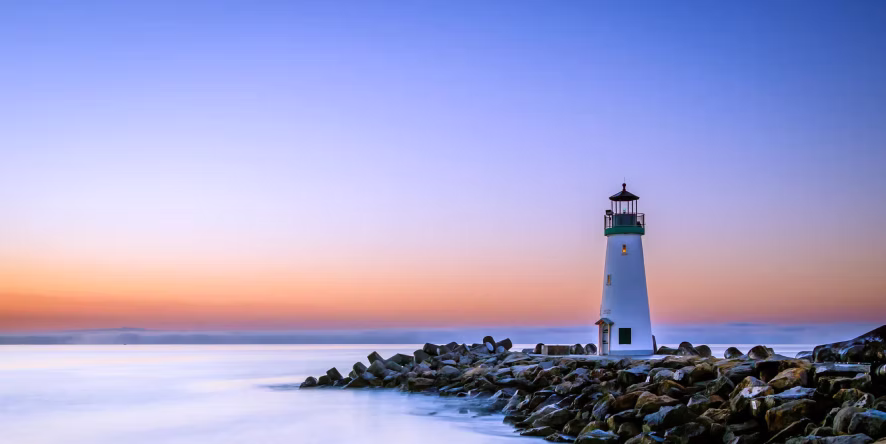 White lighthouse on rocky shore at sunset with calm water and colorful sky.