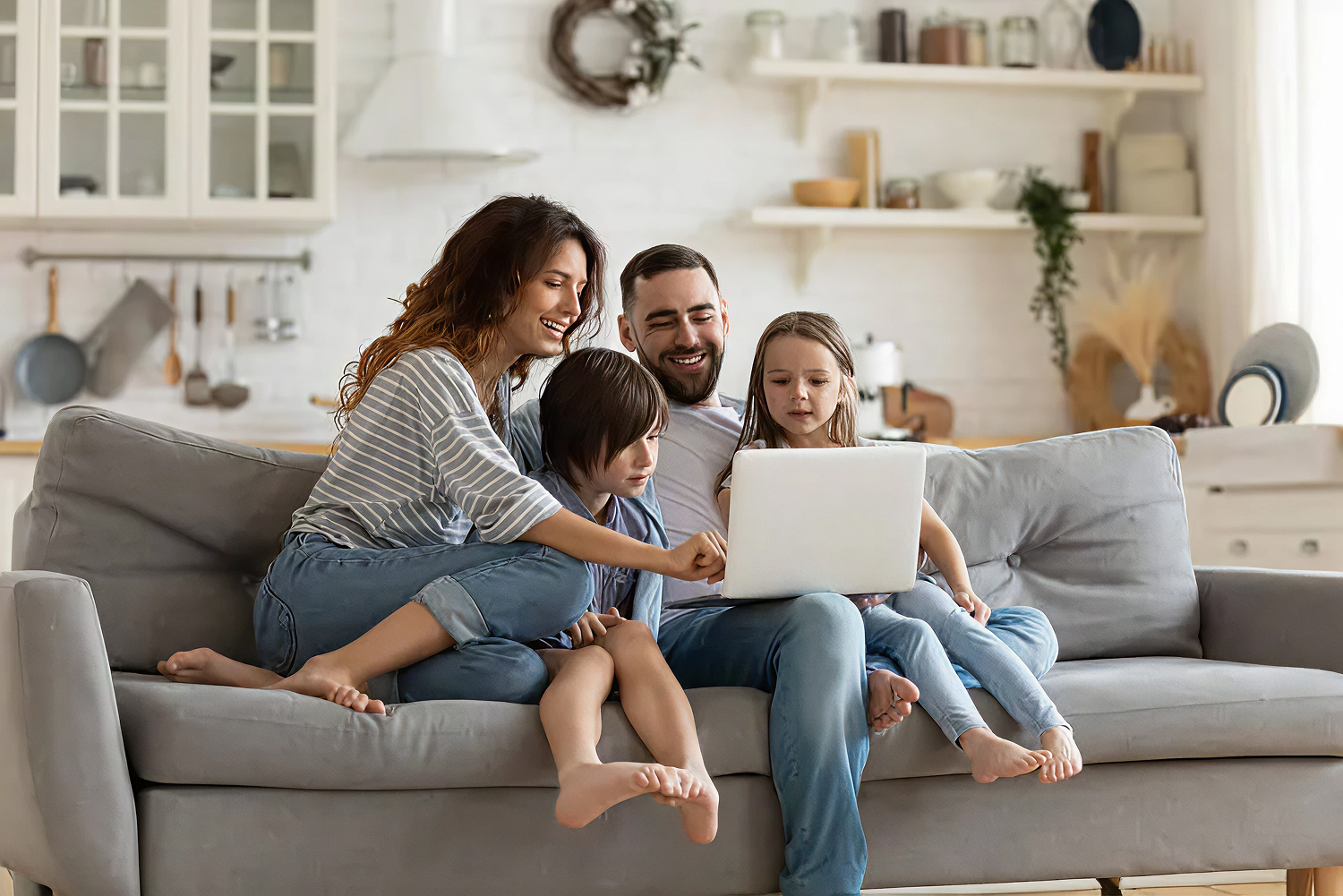 Family of four smiling at a laptop on a sofa in a cozy living room.