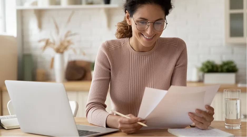 Woman reviewing documents at desk with laptop and water glass.