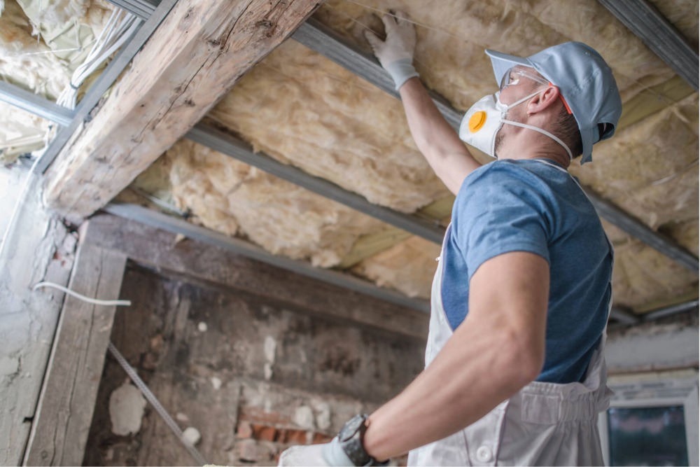 Worker installing insulation on a ceiling while wearing protective gear.