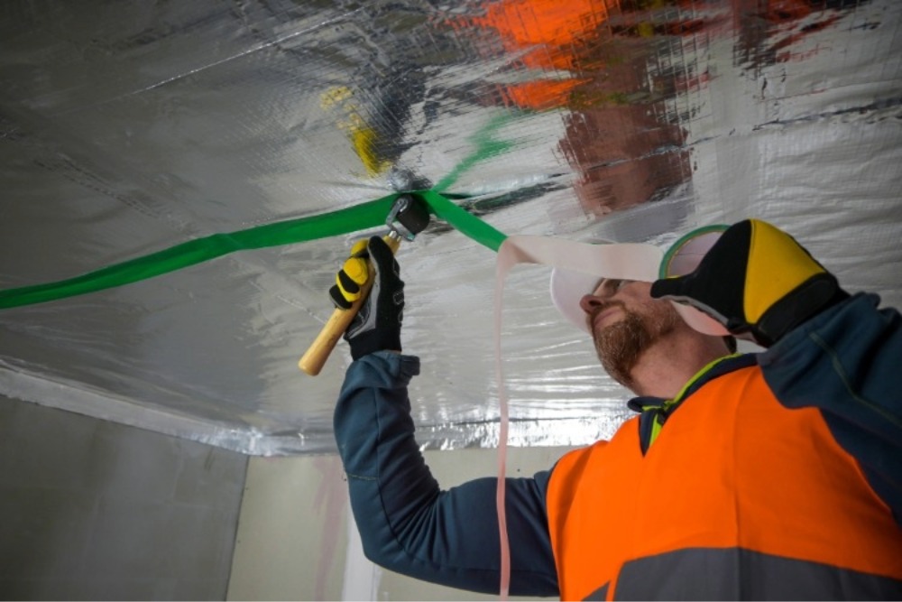 Construction worker in safety gear installs radiant barrier insulation on a ceiling using a roller and green tape.