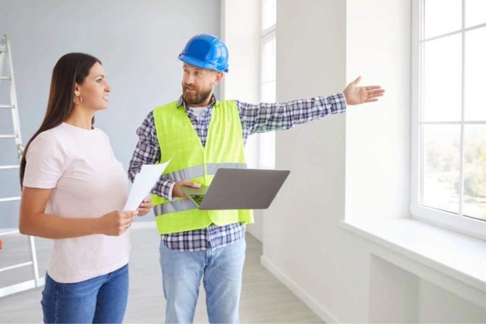 A woman reviews renovation plans with a contractor wearing a blue hard hat, while both of them hold documents.