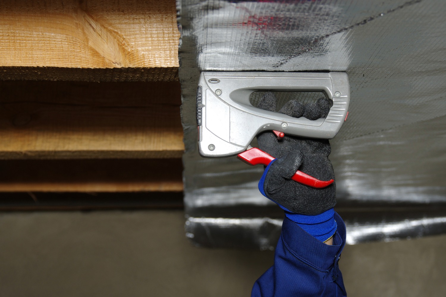 Person in a blue work uniform installing reflective radiant barrier insulation using a staple gun on wooden rafters.