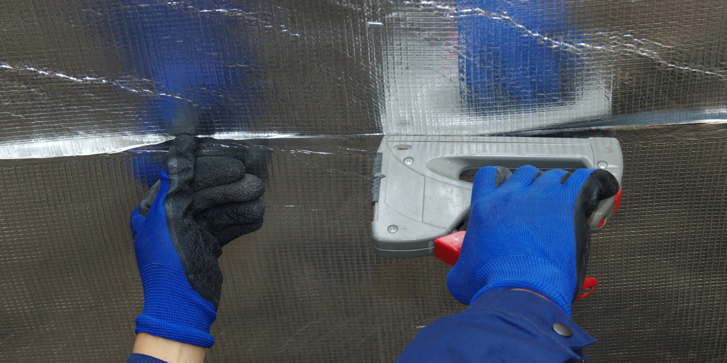 Hands in blue gloves installing radiant barrier with staple gun.