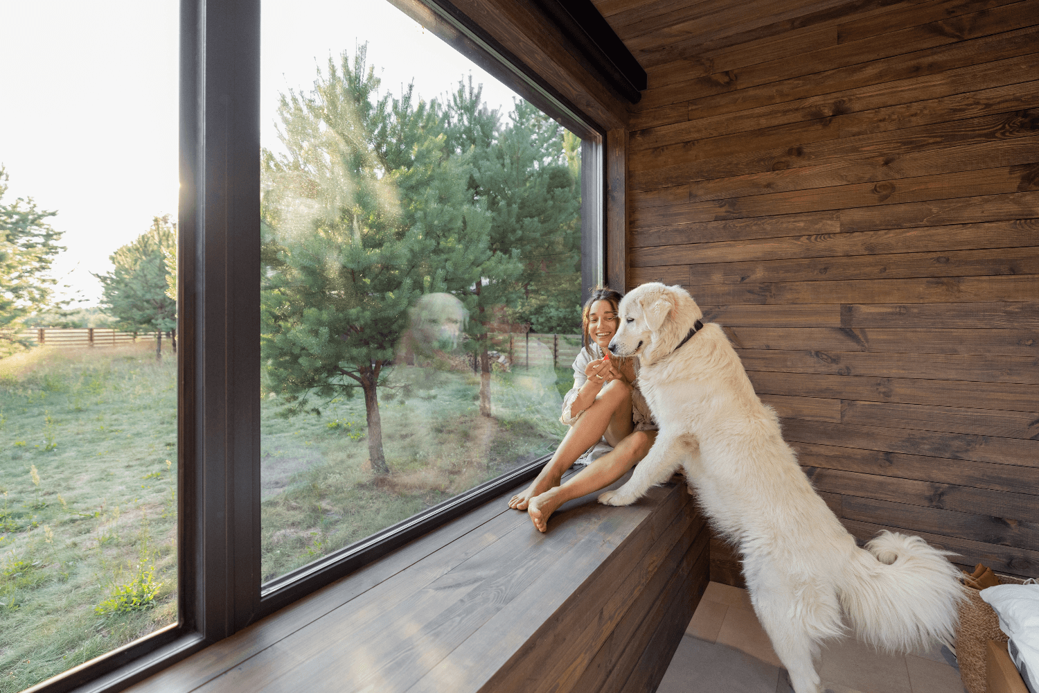 Woman with dog by large window in log cabin, forest view.