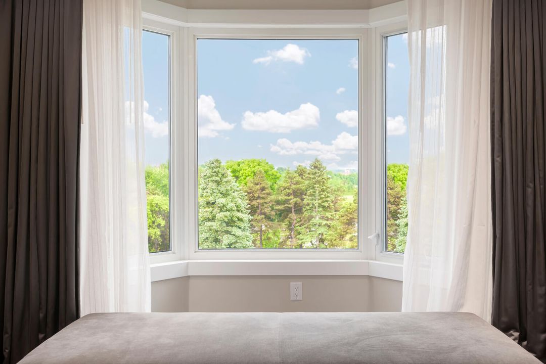 Large bay window with white curtains and tree view.